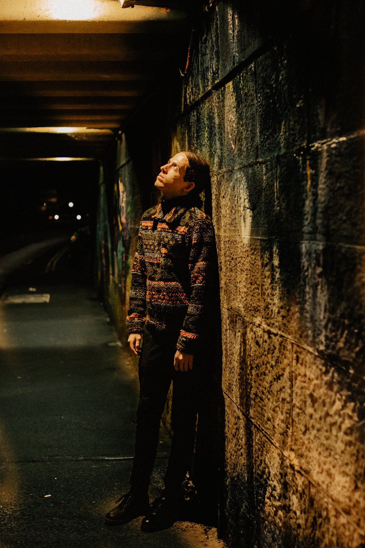 Sebastian Graysen standing against a wall under a road bridge at night, head tilted up toward a strip of light